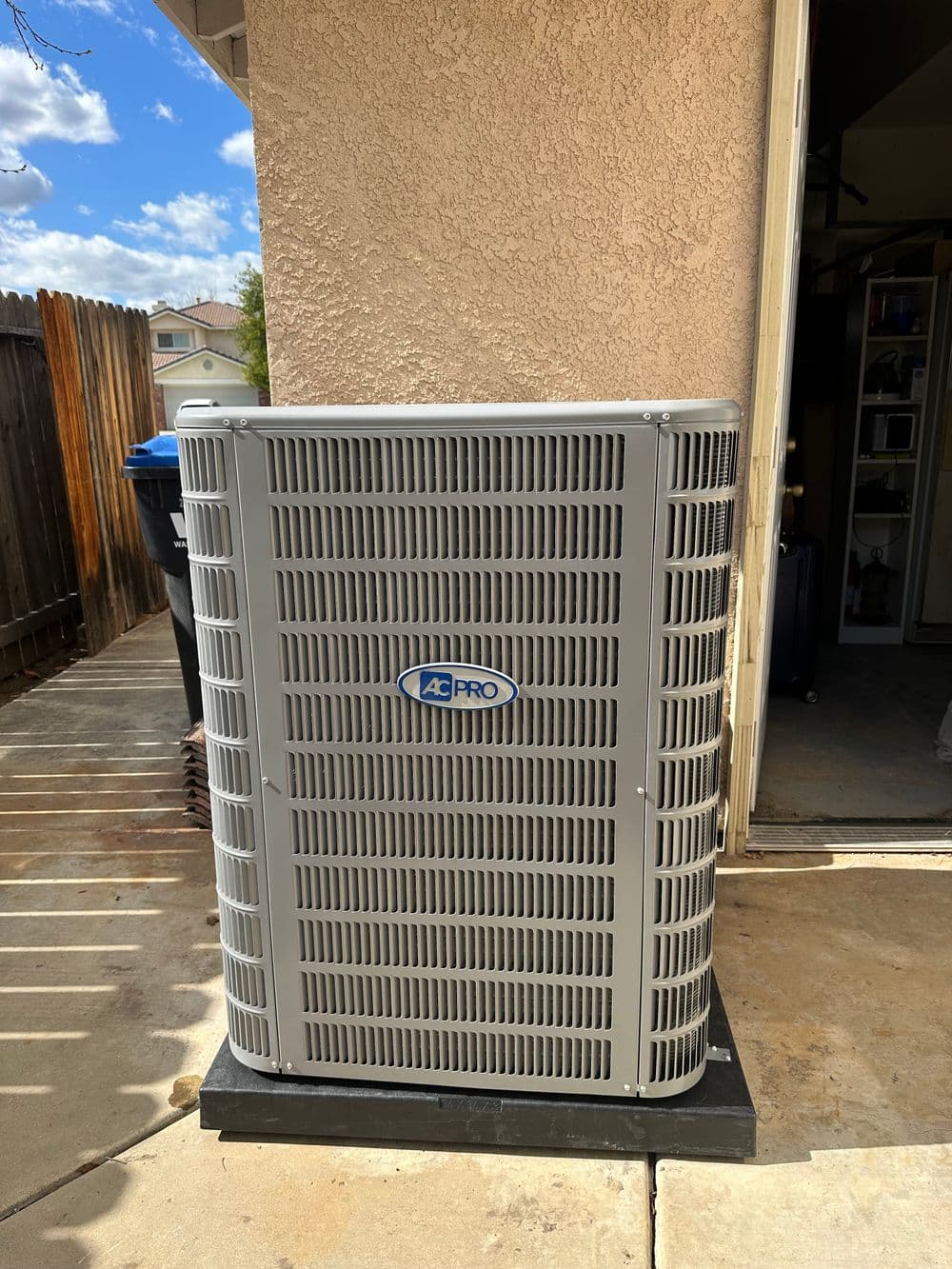 Exterior air conditioning unit on a concrete pad next to a house. Clear blue sky in background.
