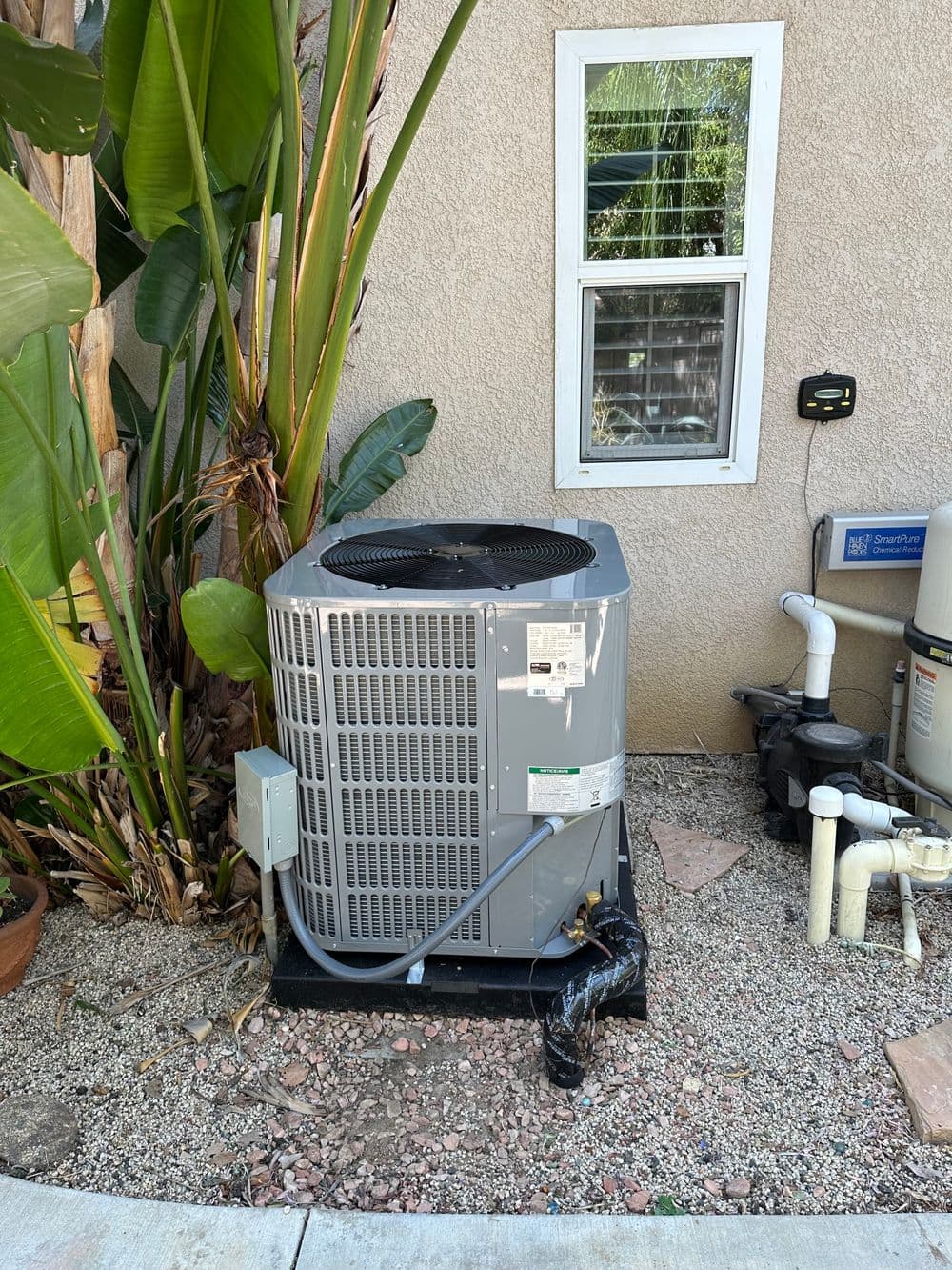 Air conditioning unit installed outside near tropical plants and plumbing equipment.