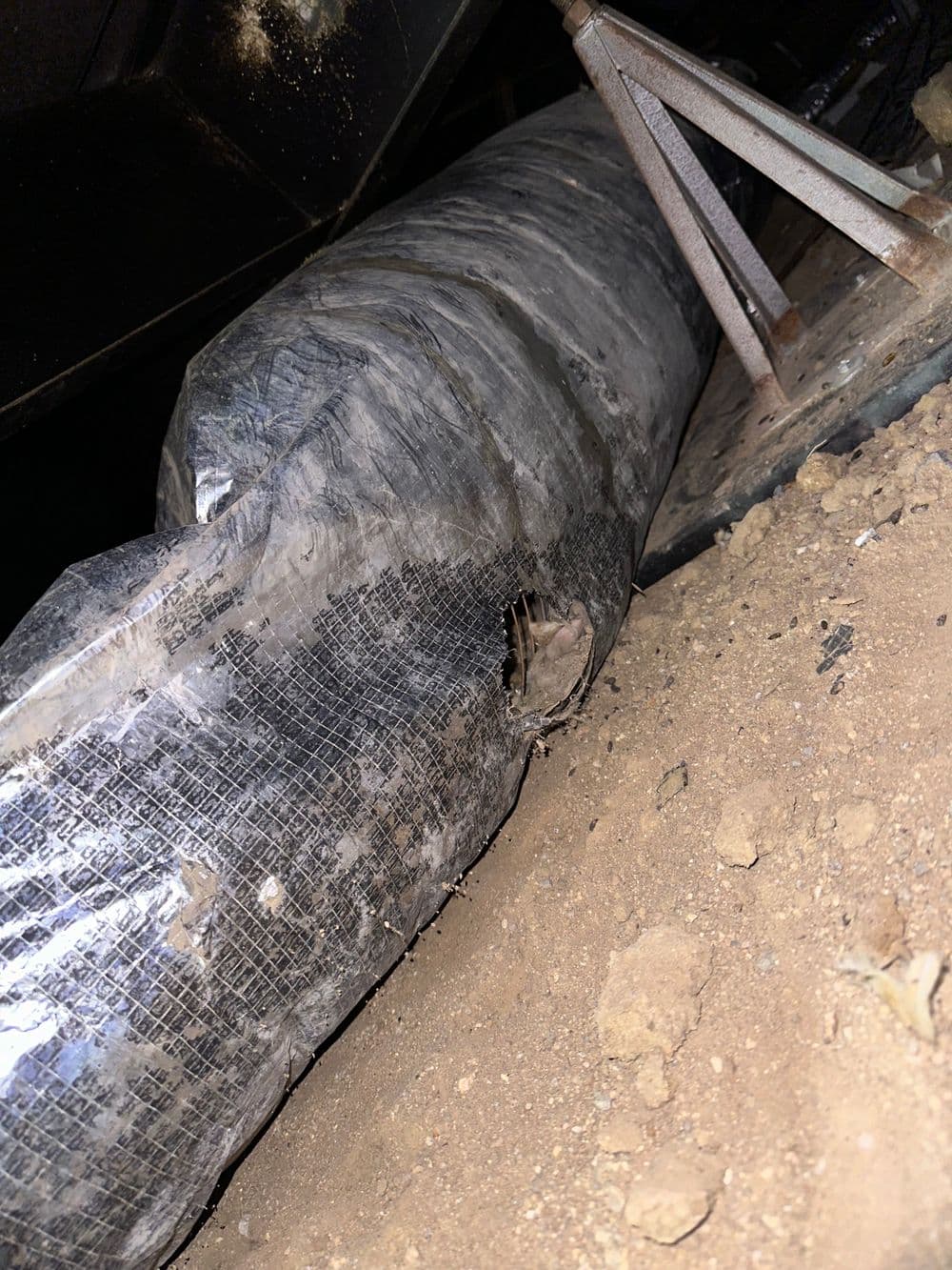 Damaged pipe wrapped in plastic, resting on dirt with support stands nearby.