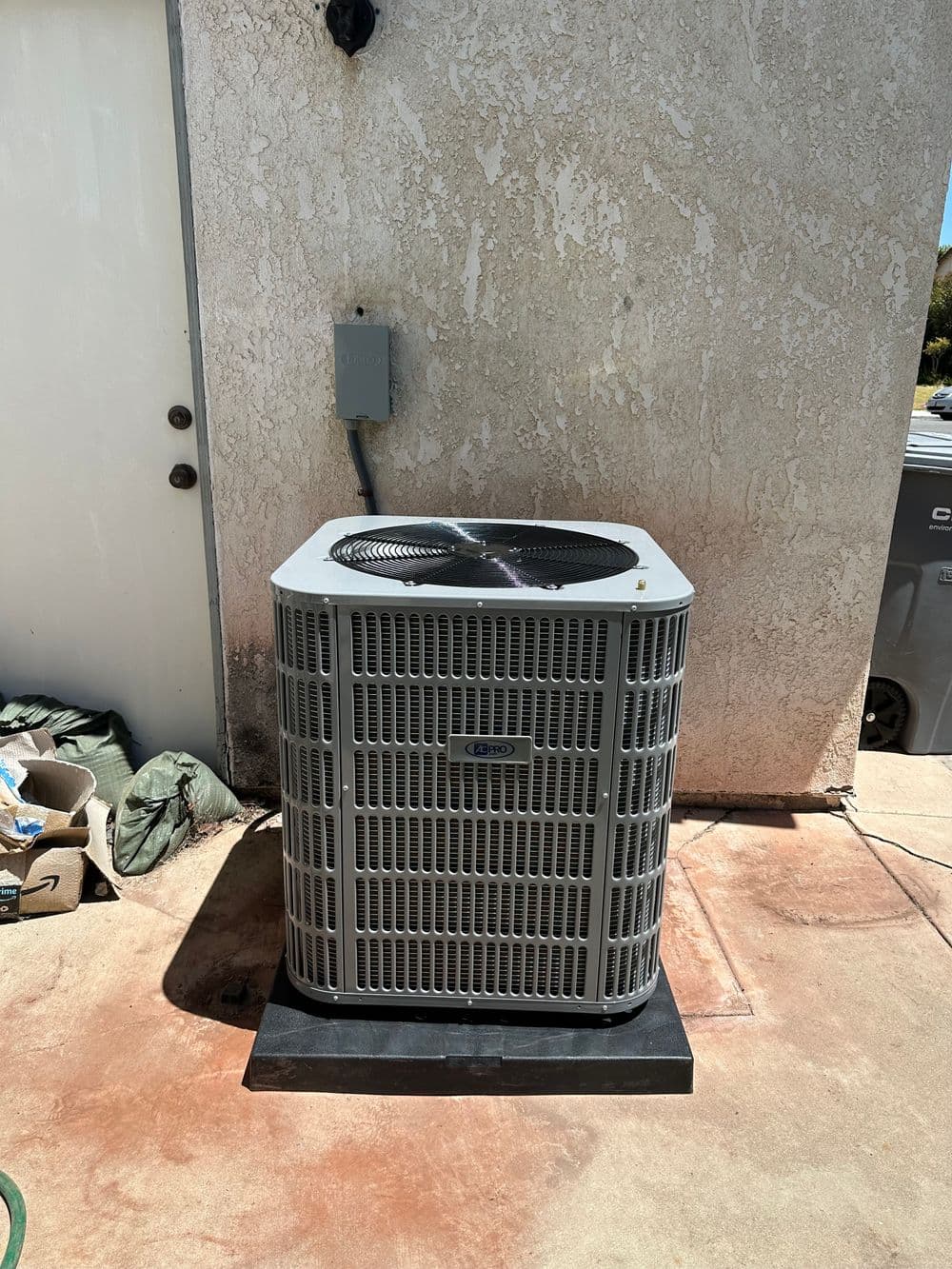 Air conditioning unit outside a house, mounted on a concrete base with nearby utility connections.
