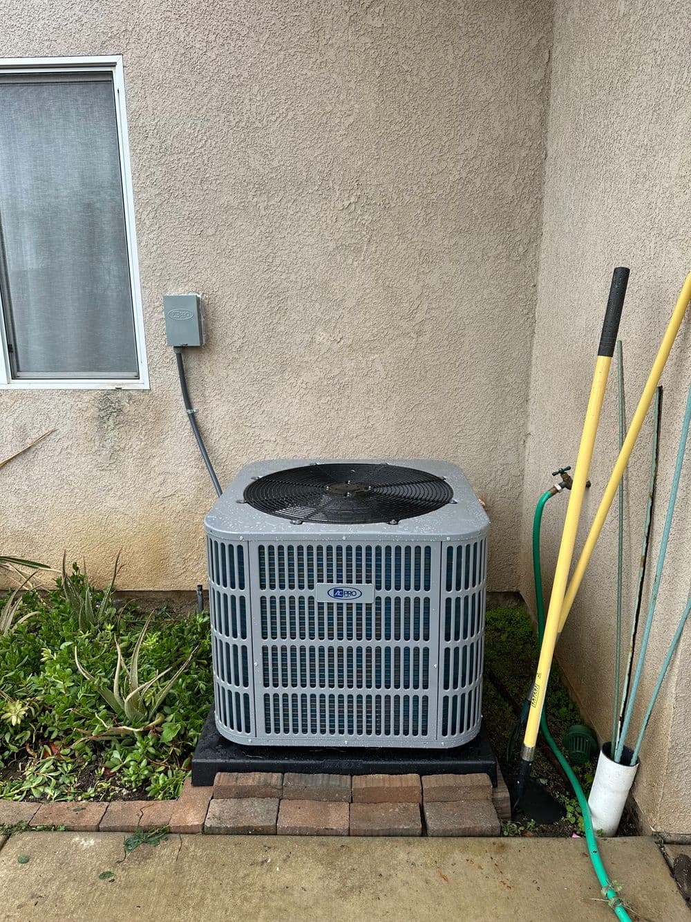 Air conditioning unit outside a house with a garden hose and landscaping around it.