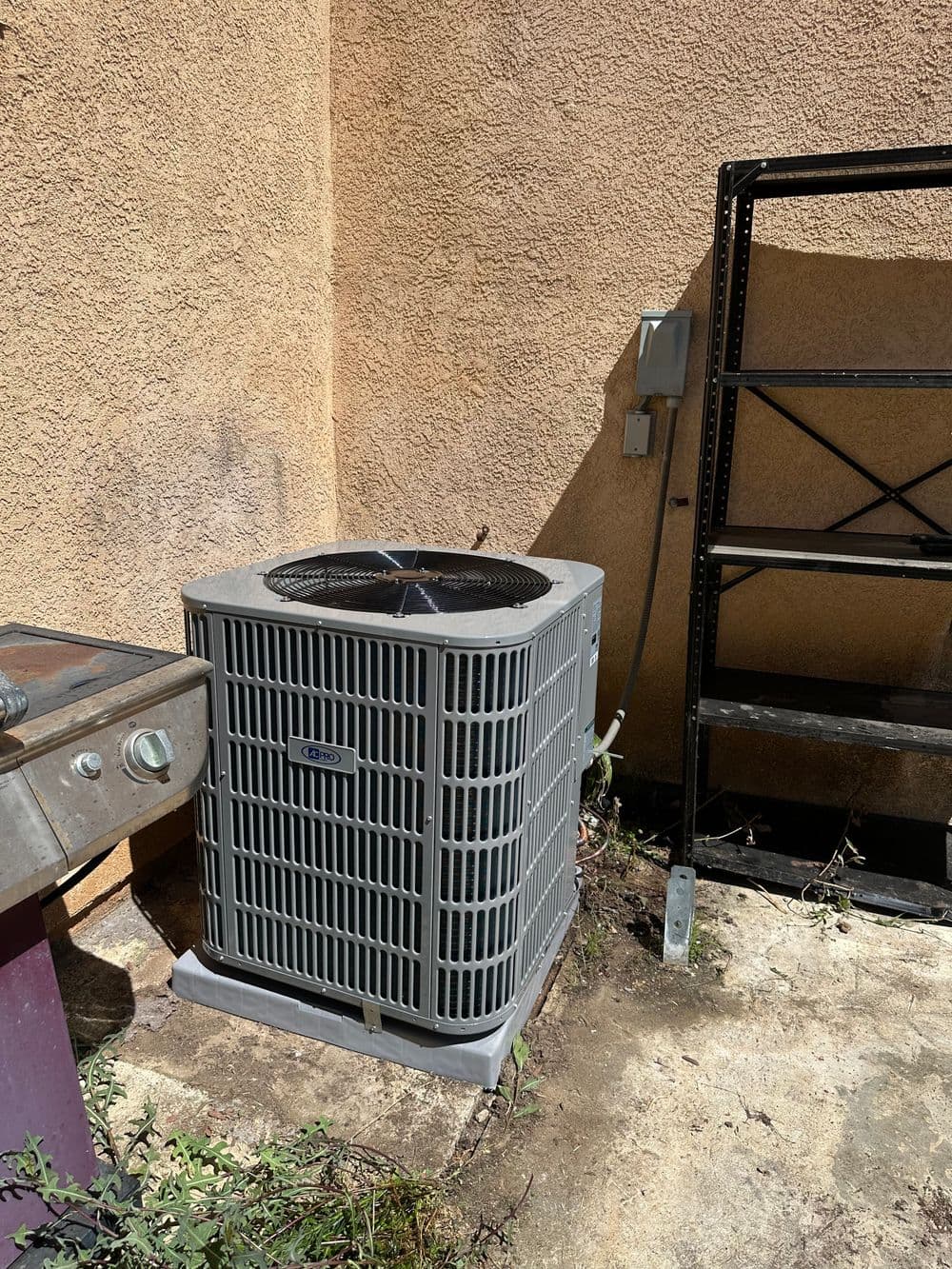 Air conditioning unit next to a grill and metal shelving against a textured wall.